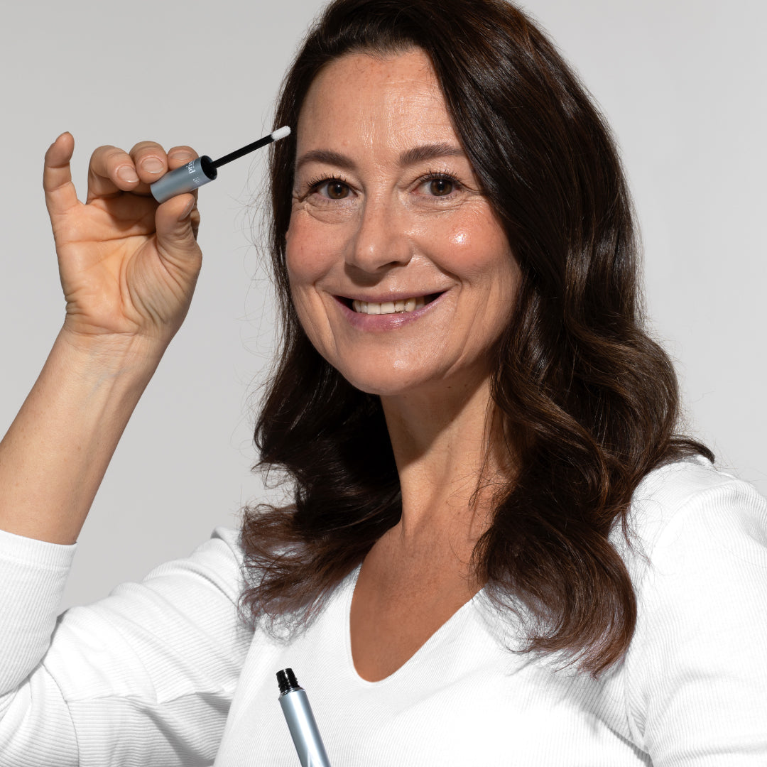 Smiling woman with long brown hair in a white top holds a mascara wand near her face, ready to apply makeup, against a plain light background.